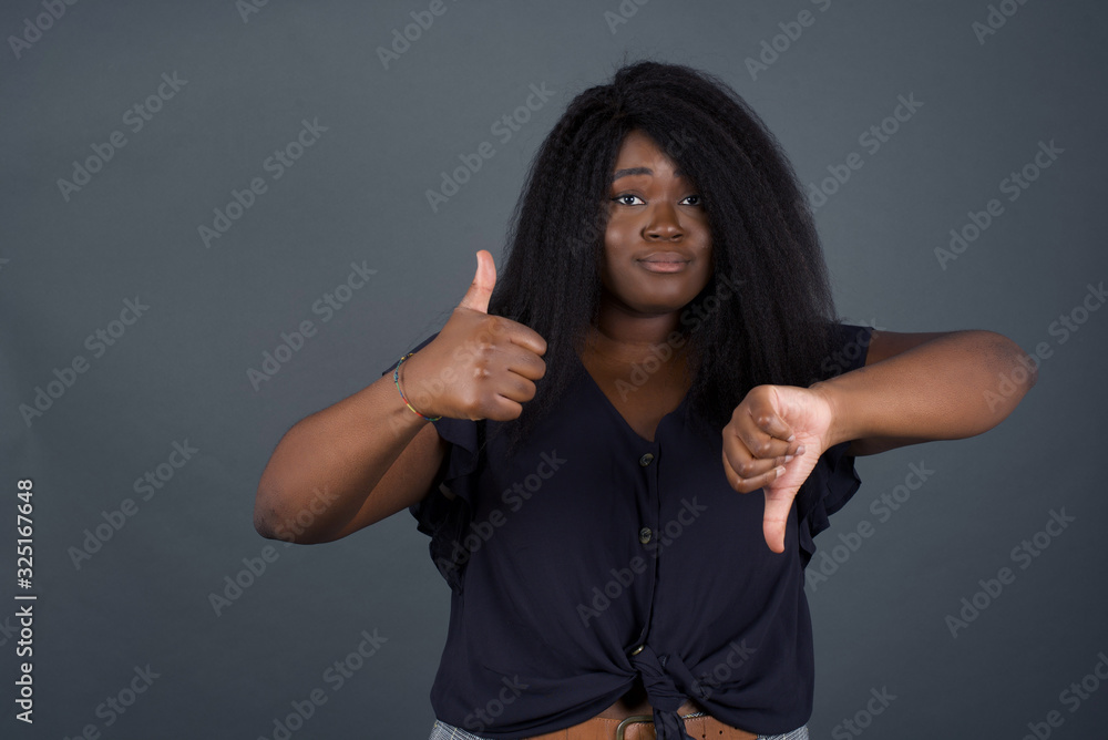 Pretty young pensive African American woman making good-bad sign. Displeased and unimpressed wearing casual clothes and standing against gray wall. Pretty young pensive African American woman making good-bad sign. Displeased and unimpressed wearing casual clothes and standing against gray wall.