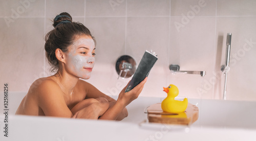 Obraz na plátně Asian girl running a bath at home taking relaxing time for herself in cozy winter apartment applying clay mask and reading a book soaking in hot water with rubber duck toy on bathtub caddy