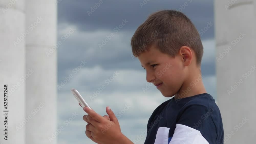 Serious boy is watching at the phone in his hands against the background of beautiful columns Serious boy is watching at the phone in his hands against the background of beautiful columns