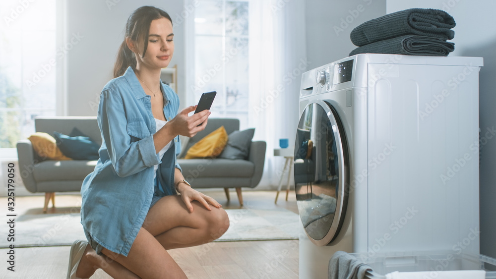 Beautiful Young Woman Sits on Her Knees Next to the Washing Machine. She Loaded the Washer with Dirty Laundry and Configured the Wash With Her Smartphone. Shot in Living Room with Modern Interior. Beautiful Young Woman Sits on Her Knees Next to the Washing Machine. She Loaded the Washer with Dirty Laundry and Configured the Wash With Her Smartphone. Shot in Living Room with Modern Interior.
