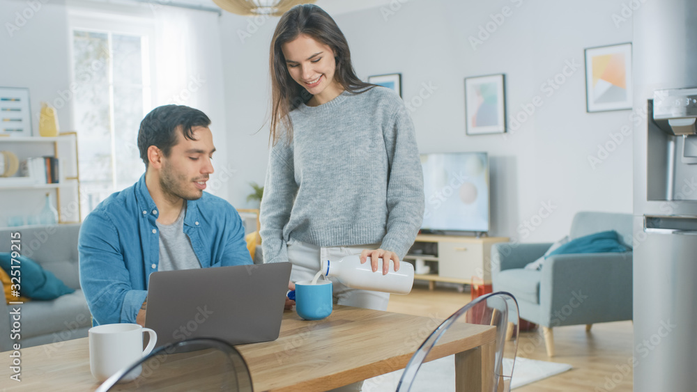 Beautiful Young Couple Talking in a Kitchen at Home. Man is Working on a Laptop, Girl d Pours Dairy Milk Yoghurt into the Mug. Thay are Happy and Laugh. Room has Modern Interior. Beautiful Young Couple Talking in a Kitchen at Home. Man is Working on a Laptop, Girl d Pours Dairy Milk Yoghurt into the Mug. Thay are Happy and Laugh. Room has Modern Interior.