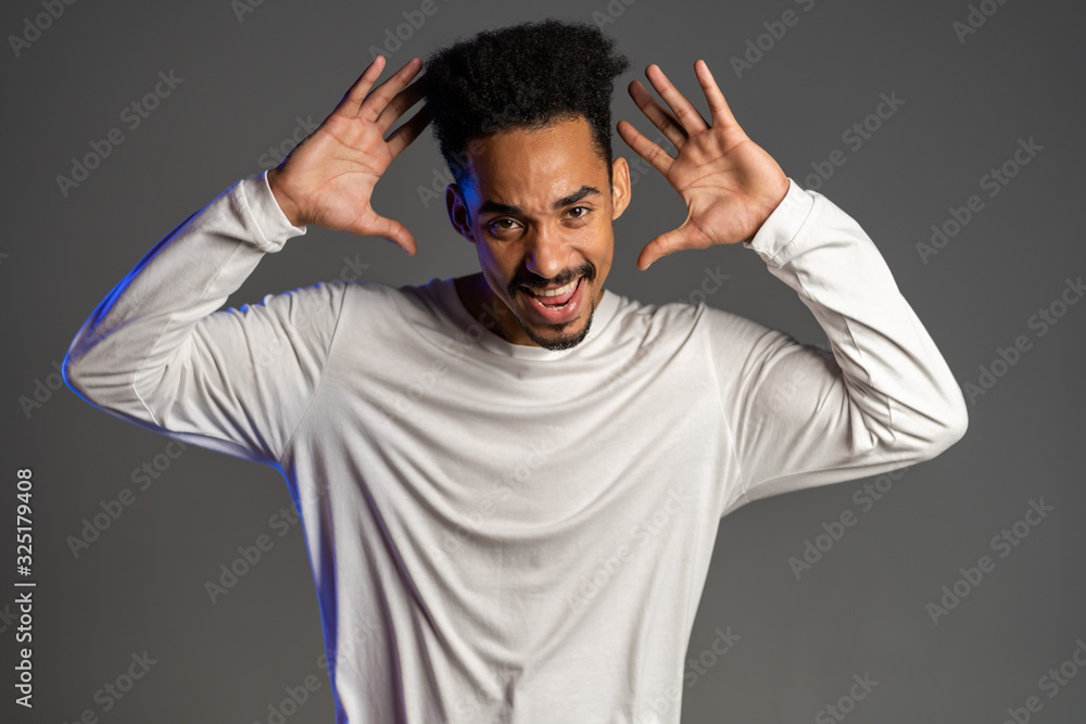 Young very active and energetic african american man in white wear smiling and dancing in good mood on grey background