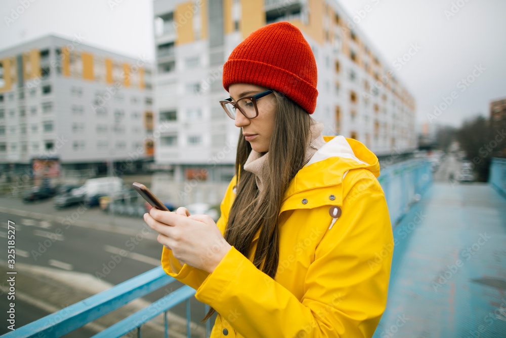 Girl in yellow raincoat checking up social media and news online. Millennial girl using mobile phone outdoors Girl in yellow raincoat checking up social media and news online. Millennial girl using mobile phone outdoors