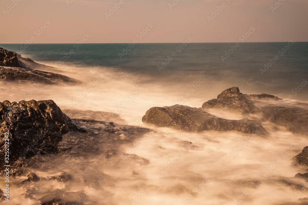 Obraz premium Beautiful rocky beach lit up by first rays of morning sunshine with golden sunlight reflecting on smooth sea water (Long Exposure Effect) 