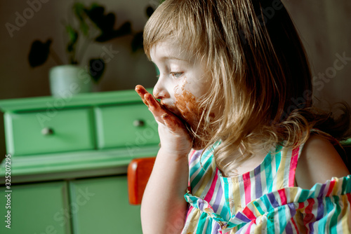 Cute curly redhead baby child shows and licks hands smeared by chocolate.Cute girl eats a bell at home. all dirty, stained with sweets. July 7 chocolate day.Indoor plants, green leaves.