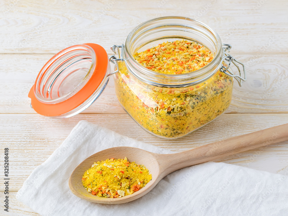 Universal seasoning in an open clip top glass jar and wooden spoon on a white wooden table. Seasoning contains salt and dried vegetables. Cooking at home concept.