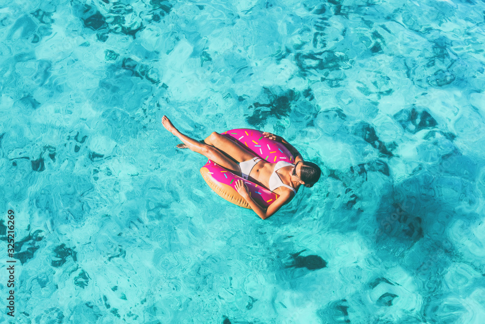 Beach vacation woman relaxing in pool float donut inflatable ring floating on turquoise ocean water background in Caribbean travel summer. Girl in white bikini top drone view.