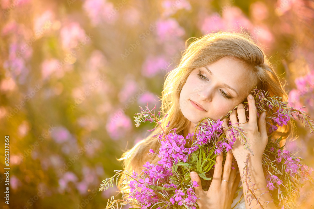 Fototapeta premium Young beautiful blonde girl gathers pink flowers in the spring blooming field at sunset.