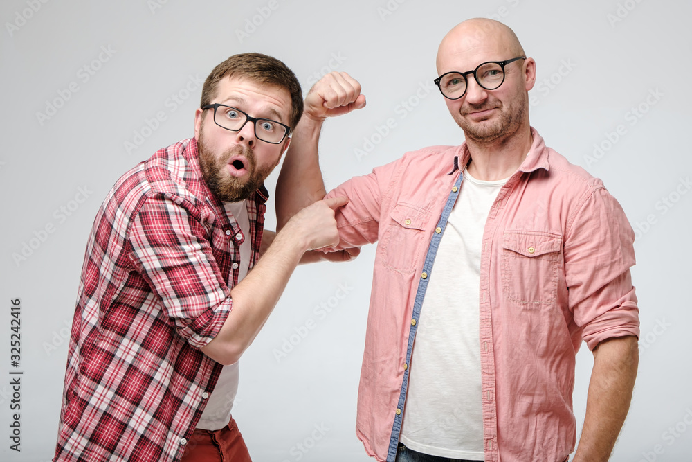 Φωτογραφία Man points his index finger at a friend biceps, he opened mouth in shock and amazement and looks into the camera with big eyes