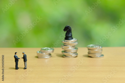 Business strategy conceptual photo - miniature businessman make handshake partnership agreement in front of coin stack with chess pawn on top of it