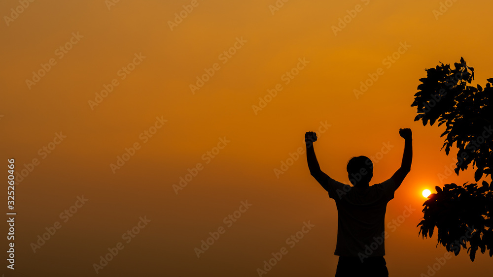 Silhouette young man with hands raised standing high up during sunset and tree background Silhouette young man with hands raised standing high up during sunset and tree background