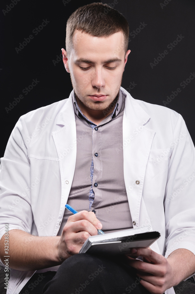 Portrait of a young intern doctor. A man sits on a chair with a notebook and pen in his hands and writes something. Making a diagnosis. Studio portrait Portrait of a young intern doctor. A man sits on a chair with a notebook and pen in his hands and writes something. Making a diagnosis. Studio portrait