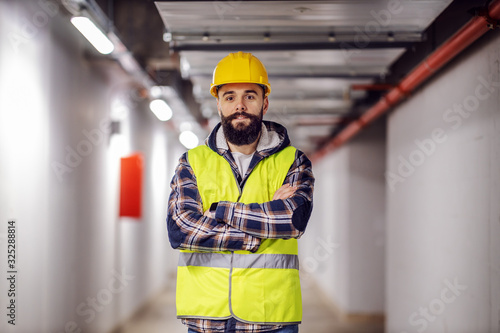 Quadro su tela Cute Caucasian bearded construction worker with safety helmet on head in vest standing with arms crossed at construction site and looking at camera