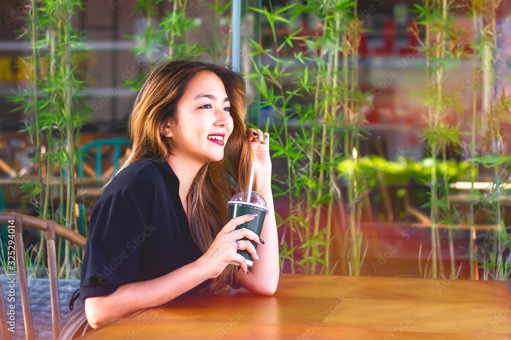 Fototapeta premium Happy, Young Asian woman drinks coffee, sits at a table in a coffee shop.Girl smiles, holds glass from drink (to go), rests on terrace 