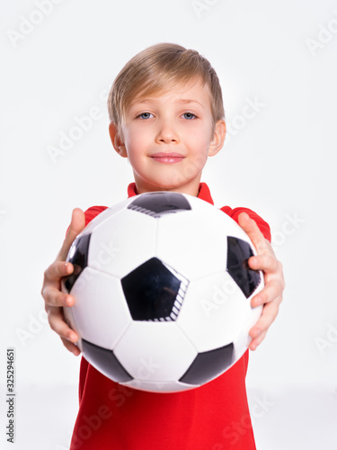 White child holds a soccer ball. Pretty 8 years old kid in a red t-shirt with a soccer ball in hand.  Photo of a boy in sportswear holding soccer ball, posing at studio. © Valua Vitaly