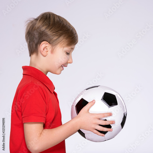 Profile portrait of a happy 8 years old kid in a red t-shirt with a soccer ball in hand. Photo of a smiling boy in sportswear holding soccer ball. White child with a smile holds a soccer ball