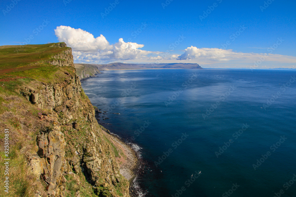 Die Landschaft einer kargen Steilküste im Norden von Island in den Westfjorden vor weitem blauen Meer mit blauem Himmel und Wolken mit Blick über die Küste