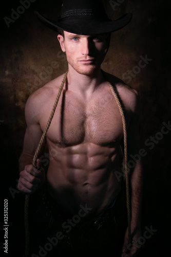 Handsome cowboy wearing a hat with barechest showing defined pecs and sixpack abs, muscular arms holding onto rope photo