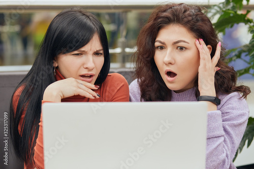 Image of surprised impressed lovely brunettes opening mouth widely, looking at laptop screen, watching shocking content, having shocked facial expression, having lunchtime. Togetherness concept. photo