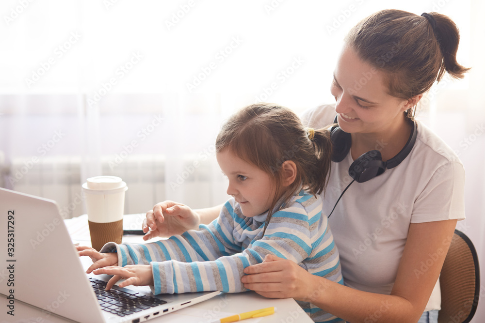 Indoor shot of young mother and child with laptop on white background, family playing computer games together, mommy wears casual white t shirt with her daughter in stripped shirt. Technology concept. Indoor shot of young mother and child with laptop on white background, family playing computer games together, mommy wears casual white t shirt with her daughter in stripped shirt. Technology concept.