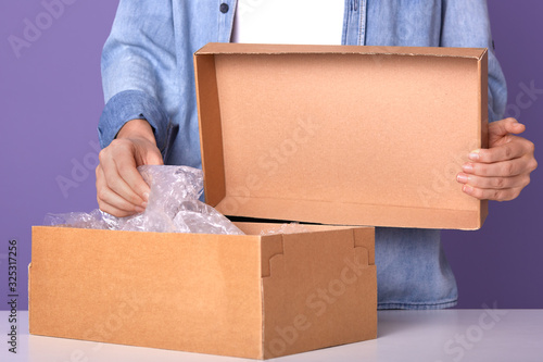 Studio shot of male hands open empty card board box isolated over lilac background, gu y wearing denim jacket and white casual shirt, man openes box with present, faceless portrait. Delivery concept photo