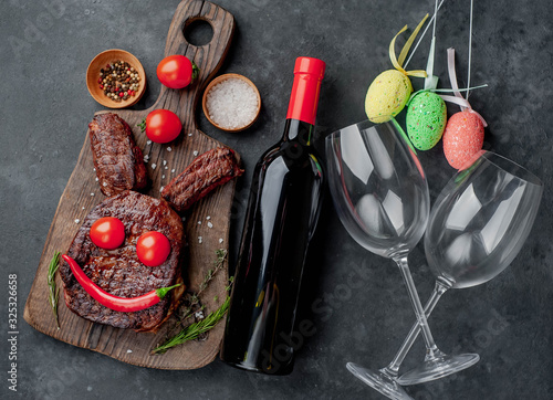 Grilled Easter steak with spices and a bottle of wine and glasses, multi-colored eggs Easter bunny on a stone background with copy space for your text.