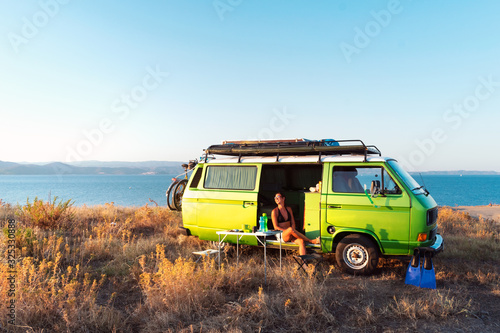 Fototapeta Young attractive female sitting in old timer camper van on a hill above the beach looking at the golden light of sunset