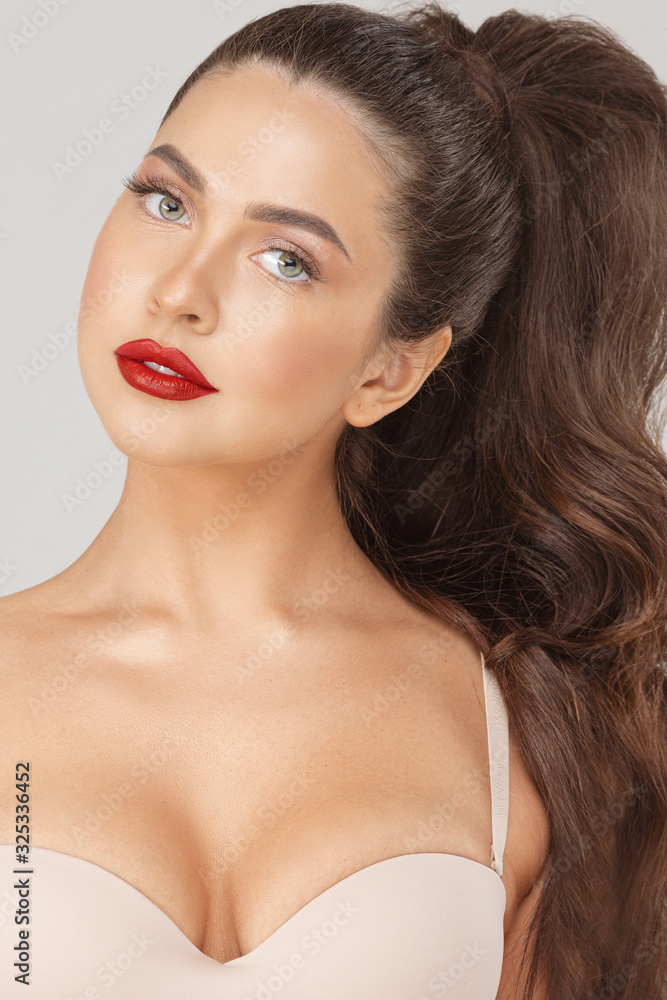 Fototapeta premium Close up portrait of attractive sexy woman with red lips posing looking at camera. Face of beauty fashionable girl in nude bra and luxury makeup isolated at white studio background