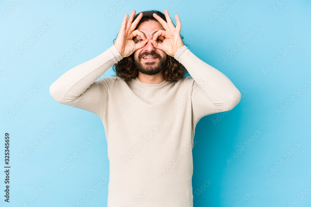 Young long hair man isolated on a blue background showing okay sign over eyes Young long hair man isolated on a blue background showing okay sign over eyes