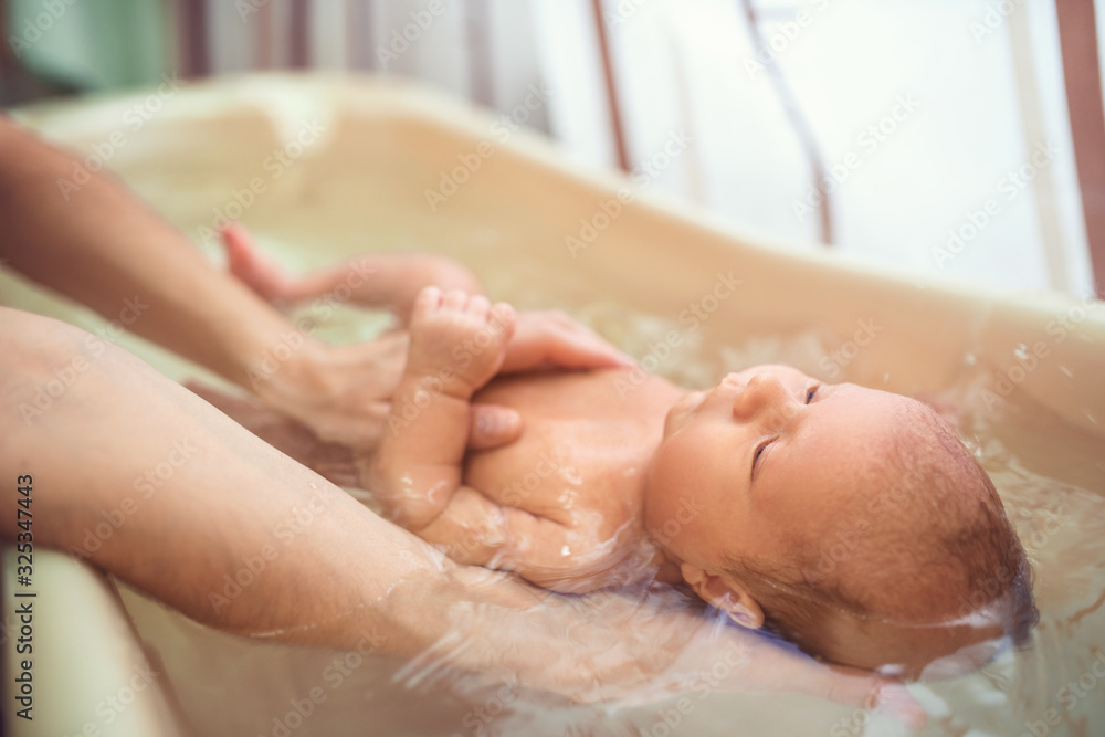 Small cute big-eyed beautiful baby bathes in warm water in the hands of a caring mother. The concept of baby care and child care - obrazy, fototapety, plakaty 