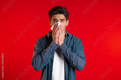 Young man sneezes into tissue. Isolated guy is sick, has a cold or allergic reaction. Coronavirus, epidemic 2020, illness concept. Red background