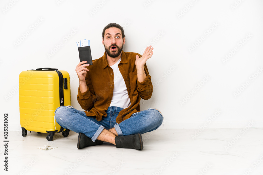 Young caucasian man sitting holding a passport and a suitcase isolated surprised and shocked. Young caucasian man sitting holding a passport and a suitcase isolated surprised and shocked.