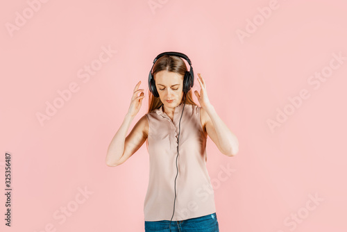 Music, leisure, hobbies. Young beautiful girl listens to music in big black headphones, pink background. Craving for art, enjoyment of creativity.