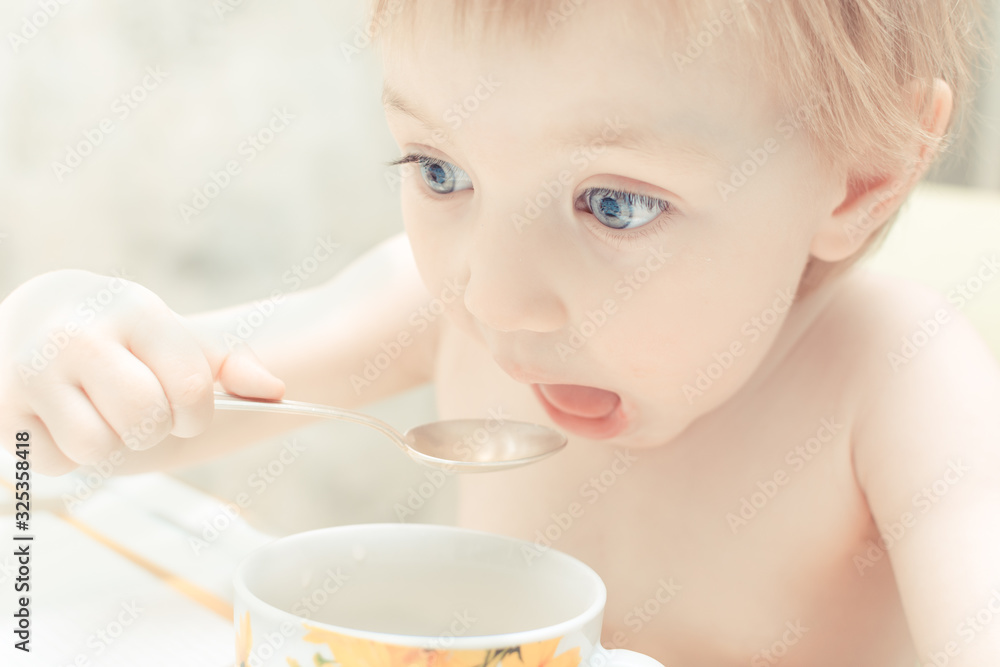 Cute child with blue eyes drinking tea from a spoon in the kitchen.