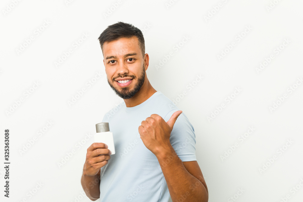 Young south-asian man holding an after shave cream points with thumb finger away, laughing and carefree. Young south-asian man holding an after shave cream points with thumb finger away, laughing and carefree.