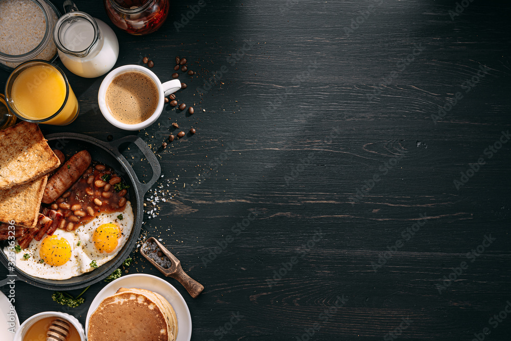 English breakfast on a dark table. fried eggs with bacon canned beans and sausage. near coffee, orange juice and pancakes with butter and honey