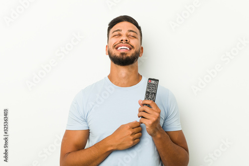 Young south-asian man holding a tv controller laughs out loudly keeping hand on chest.