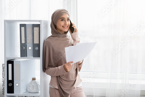 Young arabic businesswoman holding papers and talking on cellphone in office