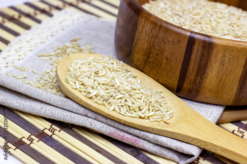 Uncooked brown rice in a bowl and spoon with light napkin on wooden background.