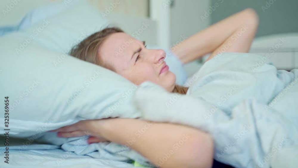 Irritated woman trying to sleep annoyed by bad noisy room neighbors covering ears with pillow, angry girl student disturbed with loud noise problem wake up lying in bed suffer from insomnia Irritated woman trying to sleep annoyed by bad noisy room neighbors covering ears with pillow, angry girl student disturbed with loud noise problem wake up lying in bed suffer from insomnia