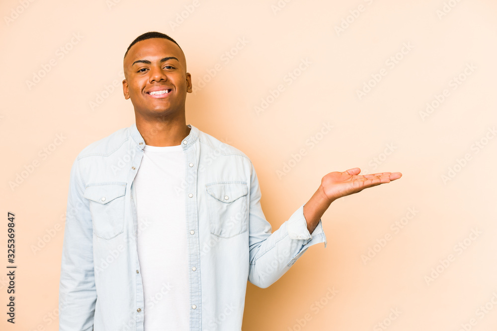 Young latin man isolated on beige background showing a copy space on a palm and holding another hand on waist. Young latin man isolated on beige background showing a copy space on a palm and holding another hand on waist.