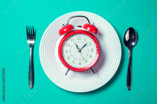 Alarm clock on the white plate with spoon and fork on the color background. Food and diet concept