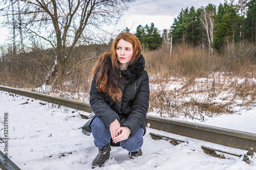 The girl with her loose hair sat on the railway and looks a little to the side against the background of the winter forest.