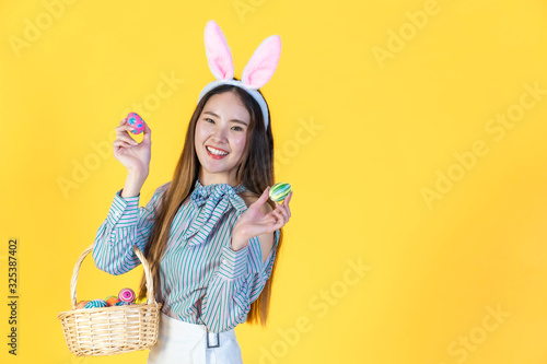 Asian young woman wearing bunny ears showing colorful easter eggs  hold basket filled eggs  toothy smiling happy face. Holidays and people concept isolated on yellow color background  copy space.