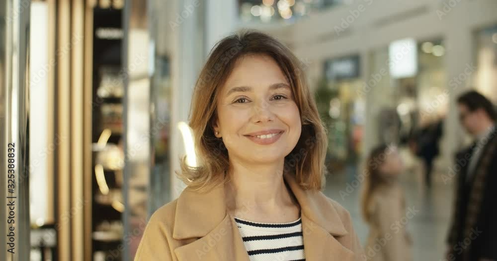 Portrait of Caucasian attractive woman in coat standing at shopping mall passage, turning face and smiling cheerfully to camera. Close up of female customer looking happy and joyful at big shop. Portrait of Caucasian attractive woman in coat standing at shopping mall passage, turning face and smiling cheerfully to camera. Close up of female customer looking happy and joyful at big shop.