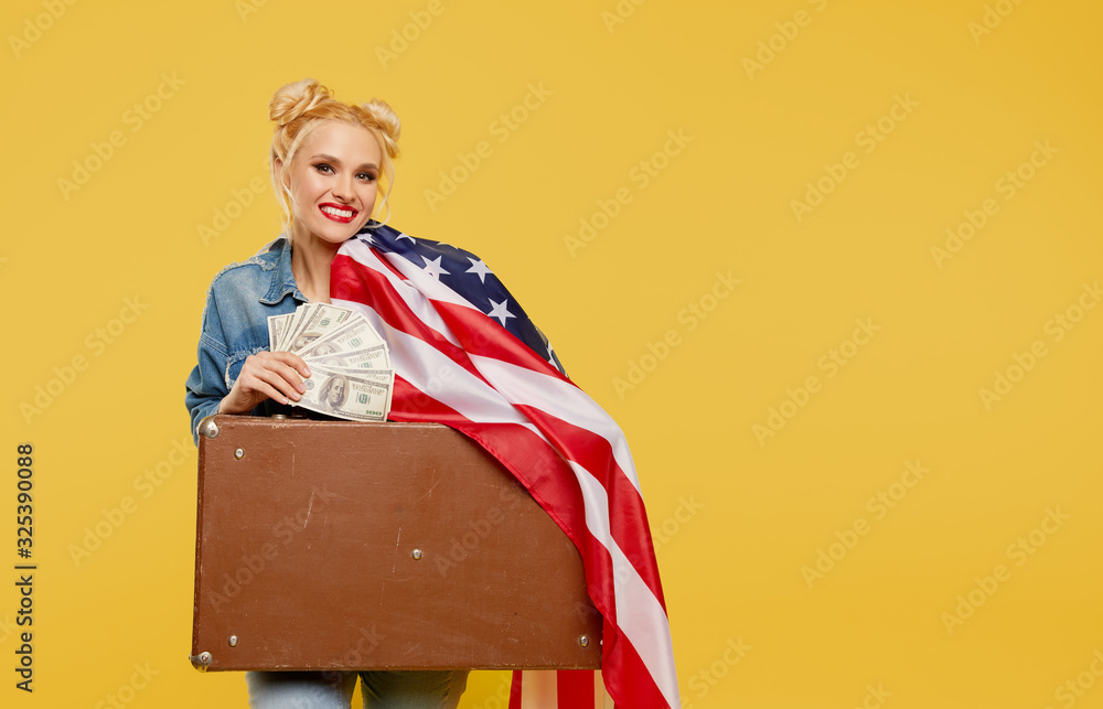 A young girl with an American flag holds in her hands a travel suitcase and banknotes of money. The concept of winning the lottery. Surprised cheerful face. A young girl with an American flag holds in her hands a travel suitcase and banknotes of money. The concept of winning the lottery. Surprised cheerful face.