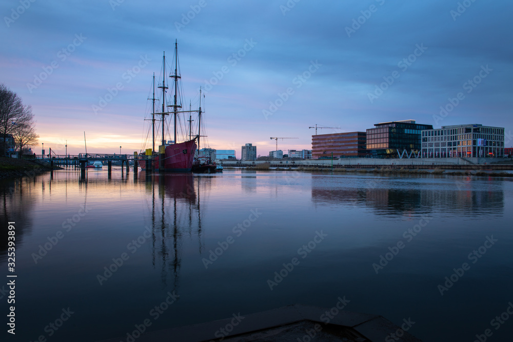 long exposure of the Überseestadt in Bremen, Germany with office building, sail boat and perfect reflection on the river weser during blue hour, last light hitting the commercial district