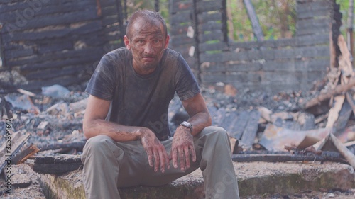 Portrait of a sad man on the background of a burned house, after fire . Consequences of fire disaster accident. Ruins after fire disaster, despair concept.
