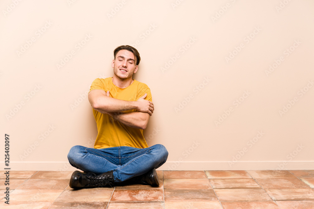 Young caucasian man sitting on the floor isolated hugs, smiling carefree and happy.