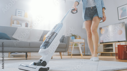 Close Up Shot of a Young Beautiful Woman in Jeans Shirt and Shorts Vacuum Cleaning a Carpet in a Bright Cozy Room at Home. She Uses a Modern Cordless Vacuum. She's Happy and Cheerful.